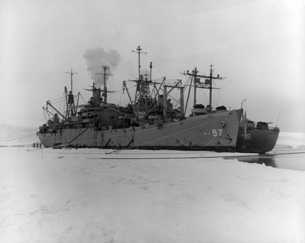 A grey naval cargo ship with tall masts and hull number 97, stopped in flat sea ice under a pale sky.