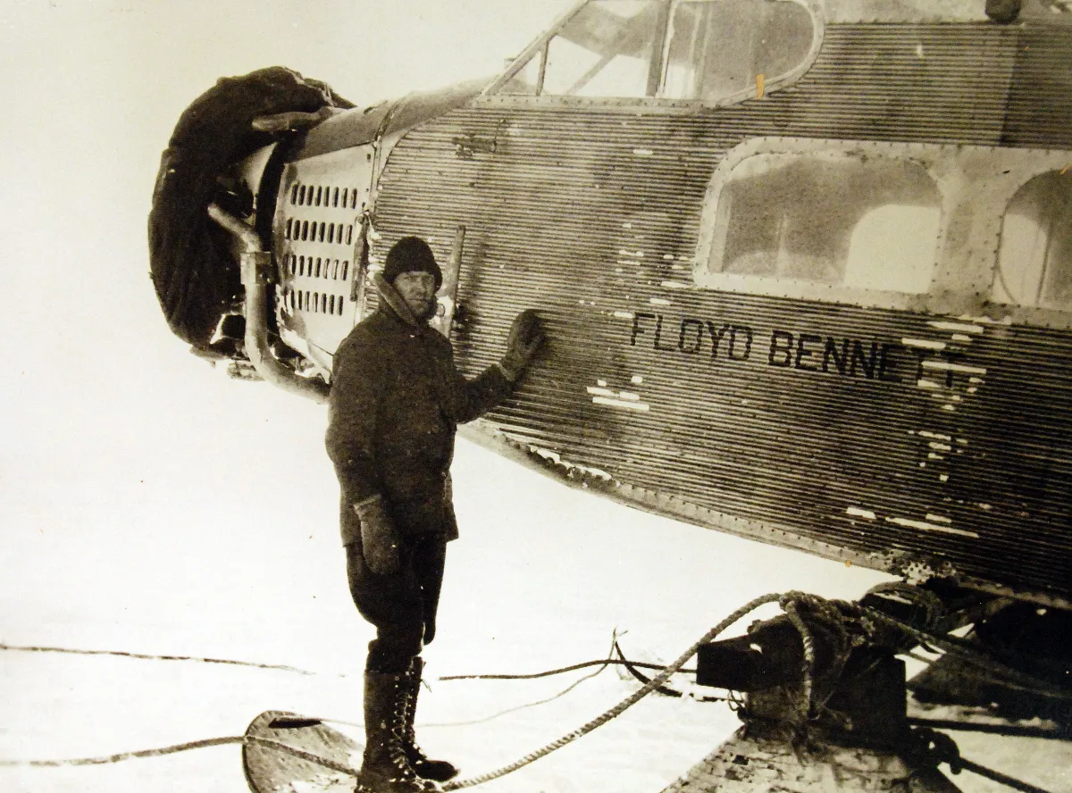A man in heavy cold-weather clothing stands beside the ribbed metal fuselage and engine of a ski-equipped aircraft lettered FLOYD BENNETT.