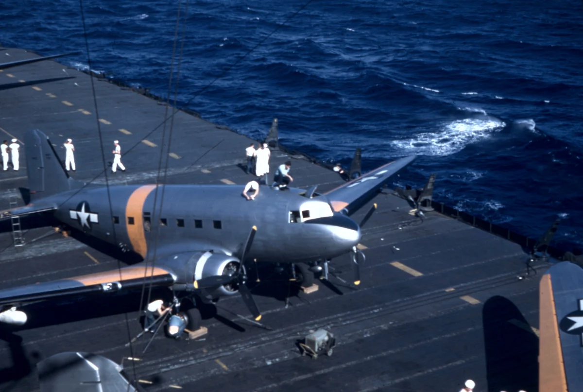 Twin-engined transport planes on skis parked on the deck of an aircraft carrier at sea, with sailors standing nearby.