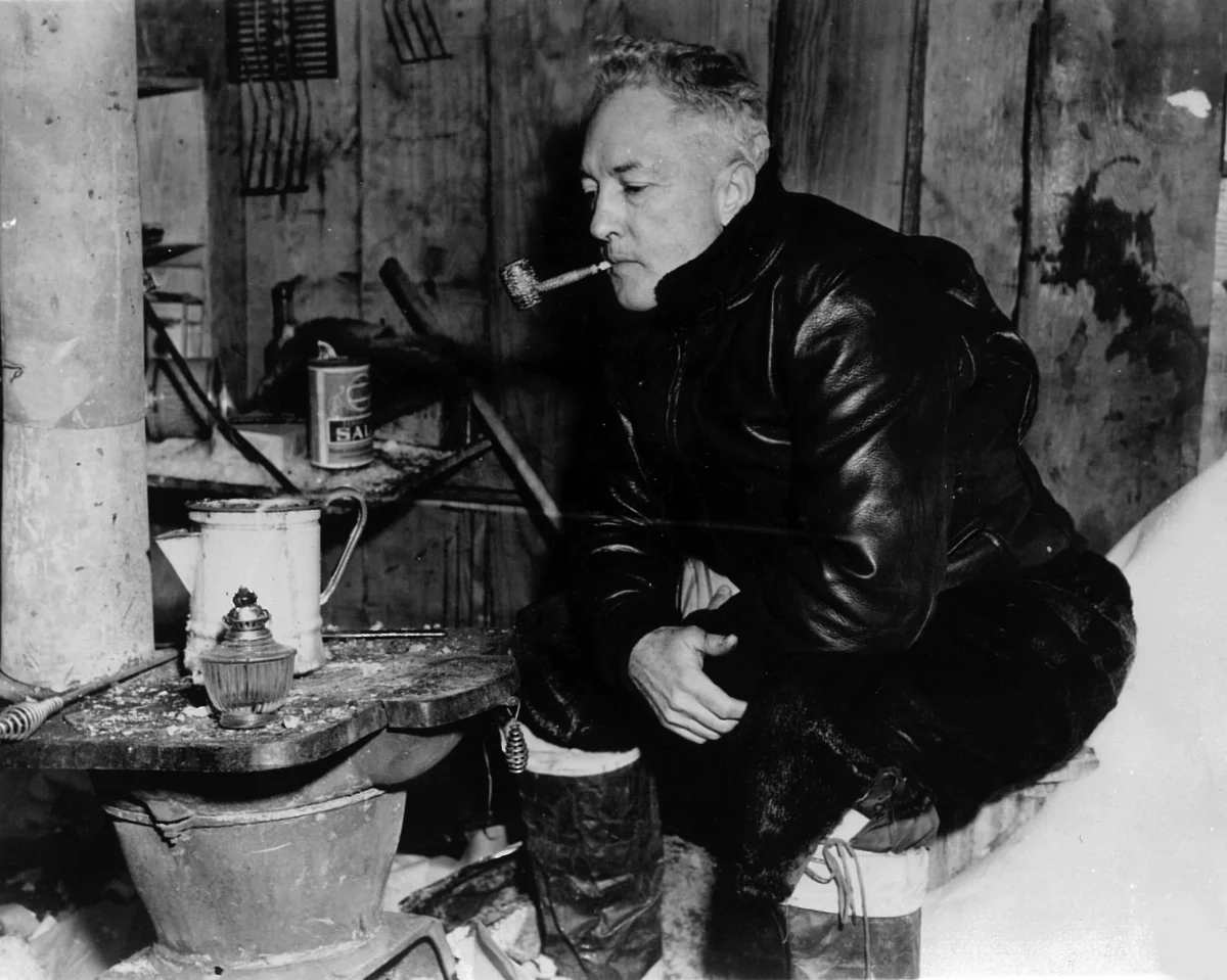 A grey-haired man in a leather flying jacket and boots crouches by a stove in a cluttered wooden hut, a pipe in his mouth.