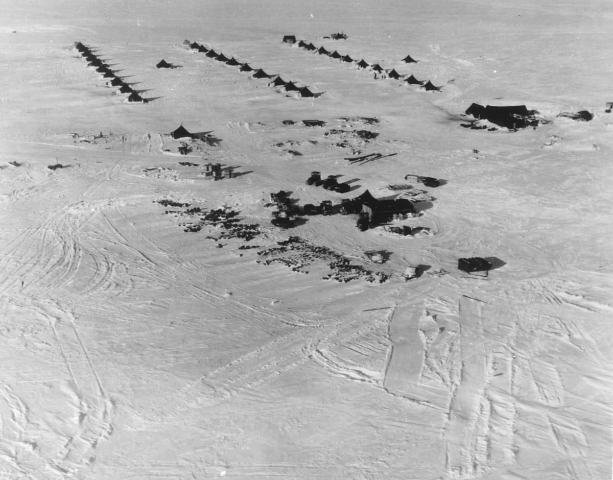 An aerial view of snow with rows of tents and dark equipment laid out in lines, and tracks crossing the surface.