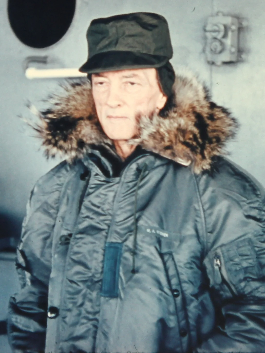 An older man in a green parka with a fur-lined hood and a dark cap stands on the deck of a ship, in colour.