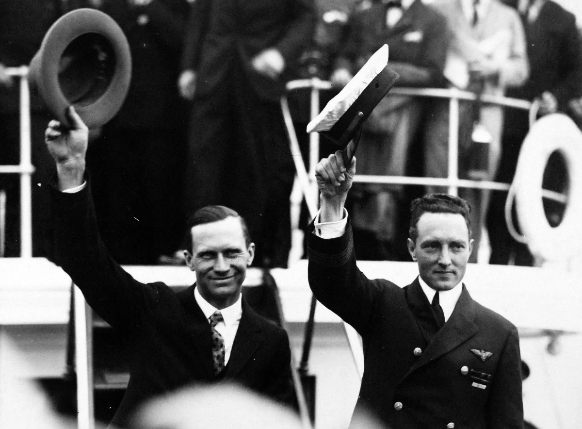 Two men in dark suits on a ship's deck raise their hats to a crowd.