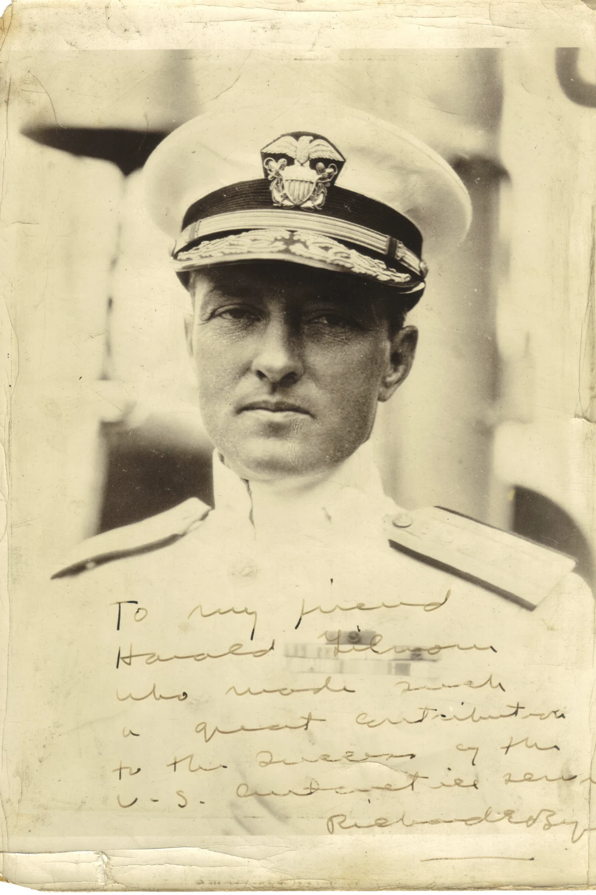 A studio portrait of a naval officer in a white uniform and peaked cap, with a handwritten dedication in ink across the lower part of the stained print.