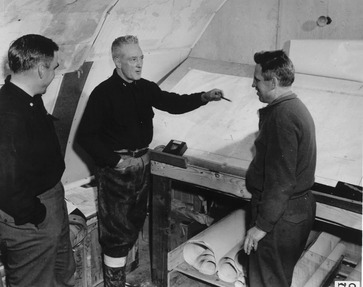 A black and white photograph of three men in a hut, one pointing at a chart pinned to the wall while the other two look on.