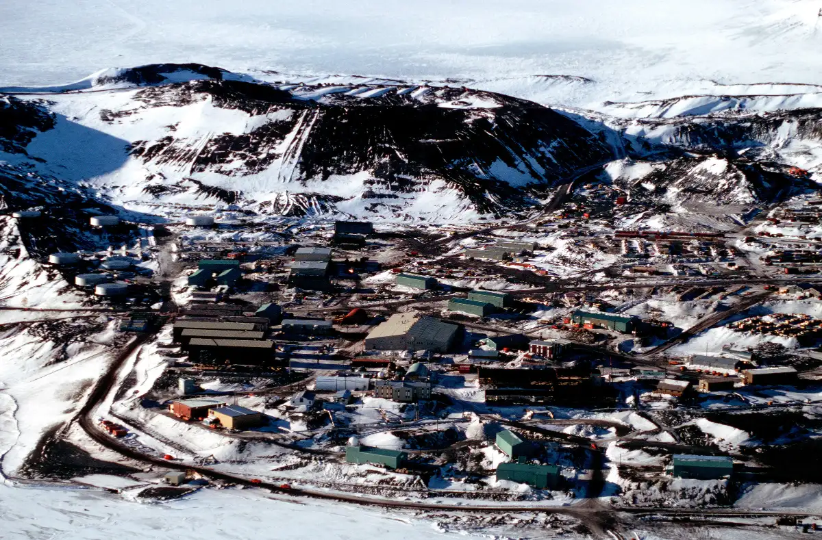 An aerial view of a scientific station: low buildings, roads and tanks among snow and dark rock below a hill.