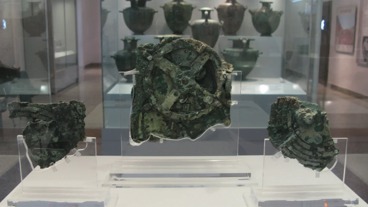 Three corroded bronze fragments on clear stands in a museum case, the largest in the middle with a spoked wheel, bronze water jars on shelves behind.
