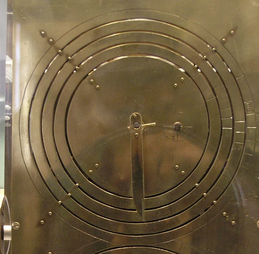 The brass back plate of a replica of the mechanism, with an upper dial of several concentric rings and a pointer.