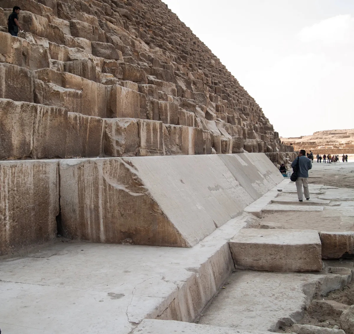 Pale smooth casing blocks still in place at the foot of the Great Pyramid, with the rough stepped core rising behind them and a visitor for scale.