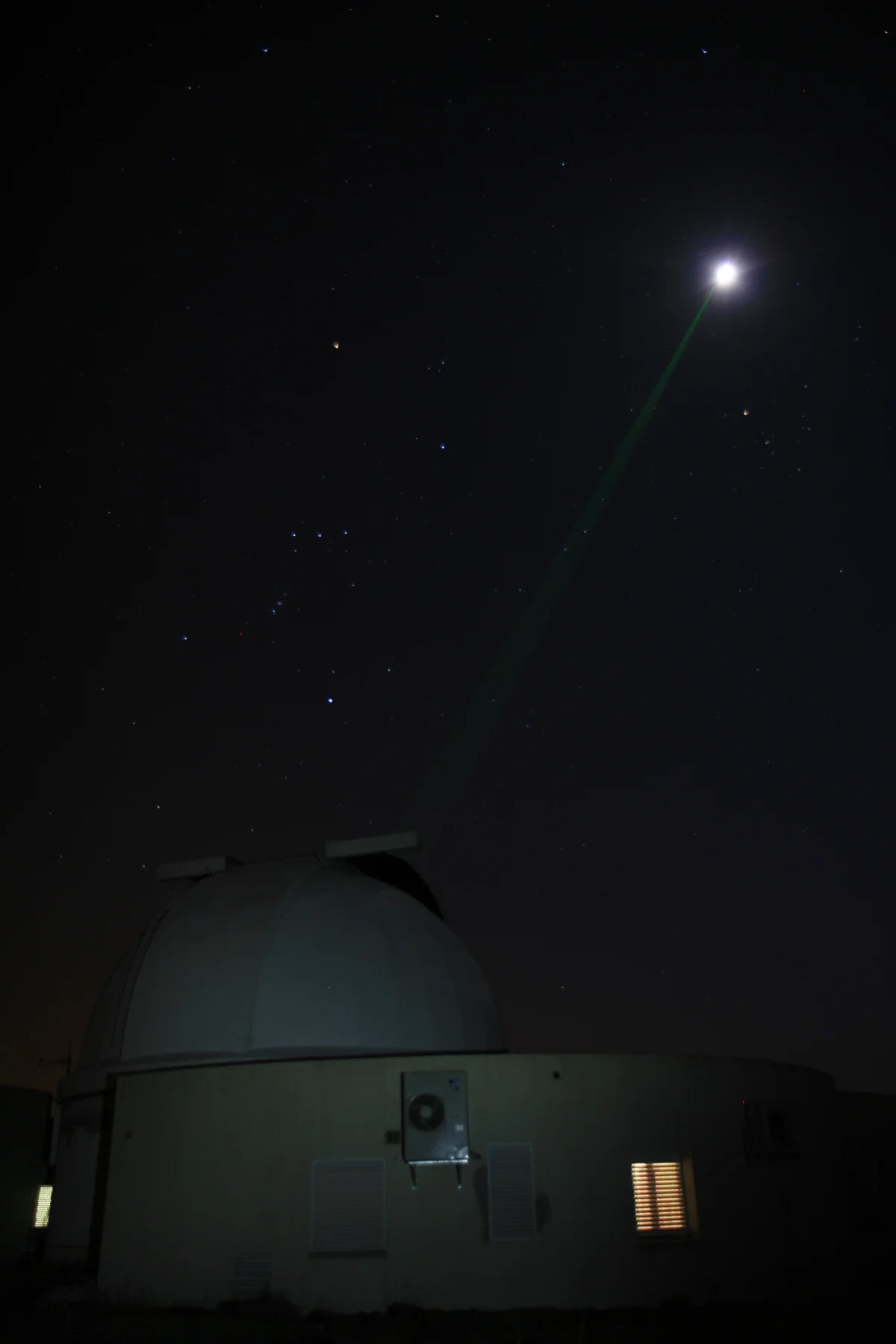 A thin green laser beam rises from an observatory dome at the bottom of the frame to the bright Moon at upper right, across a dark starry sky.
