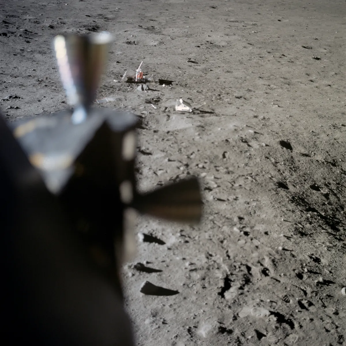 A blurred thruster of the lunar module fills the left of the frame; grey ground runs out to a small copper instrument and a white box in the distance.