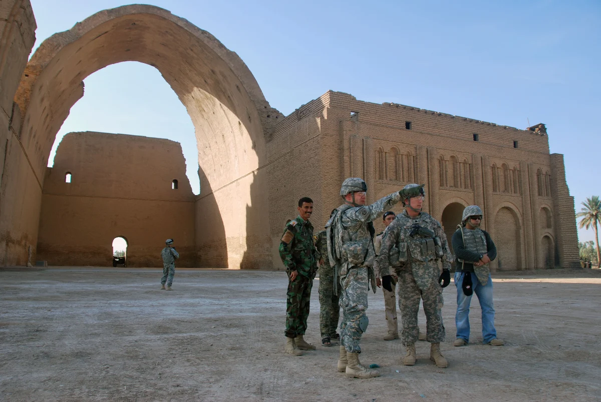 The great brick arch of Ctesiphon and its ruined wall on a flat plain, with a group of uniformed soldiers standing in the foreground.
