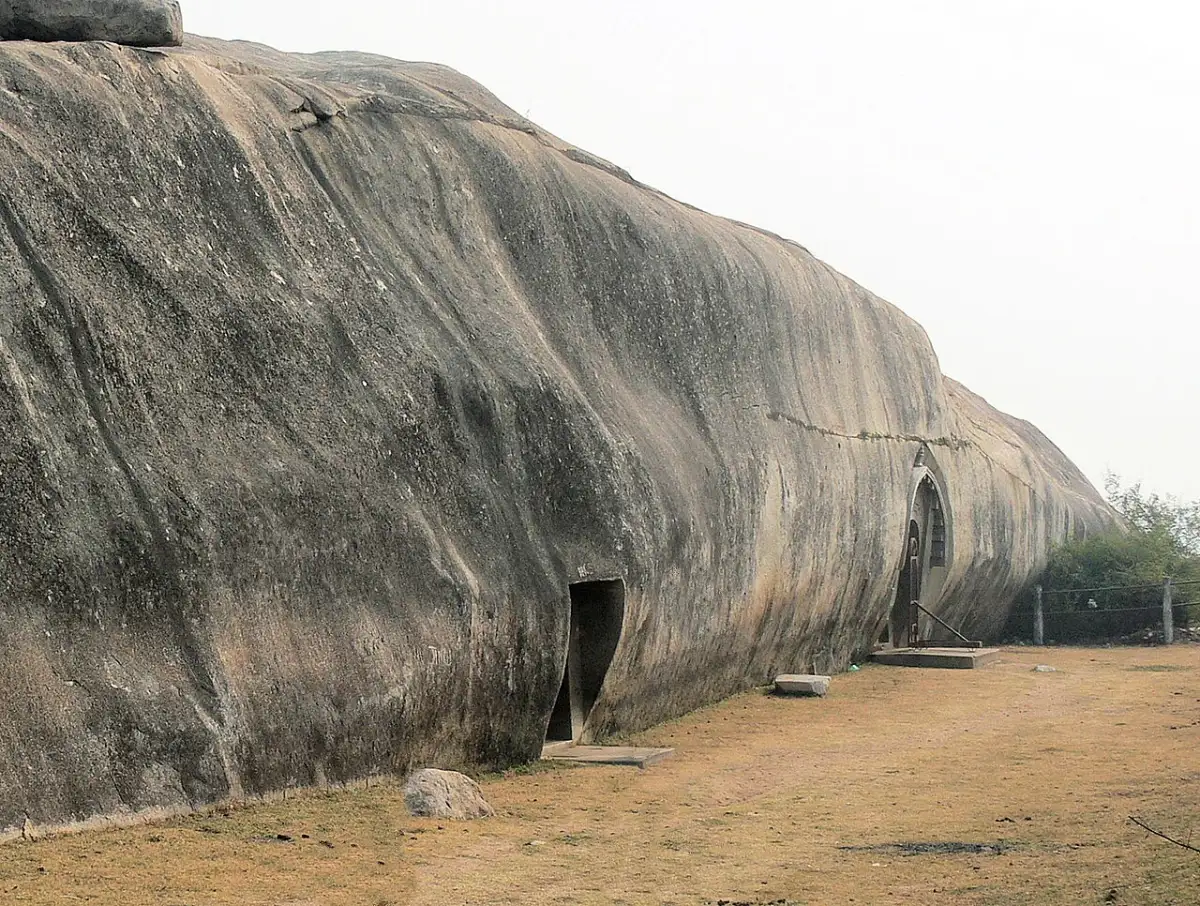 A long bare whaleback of grey granite with two doorways at its foot, one plain and one arched, and a dirt path beside it.