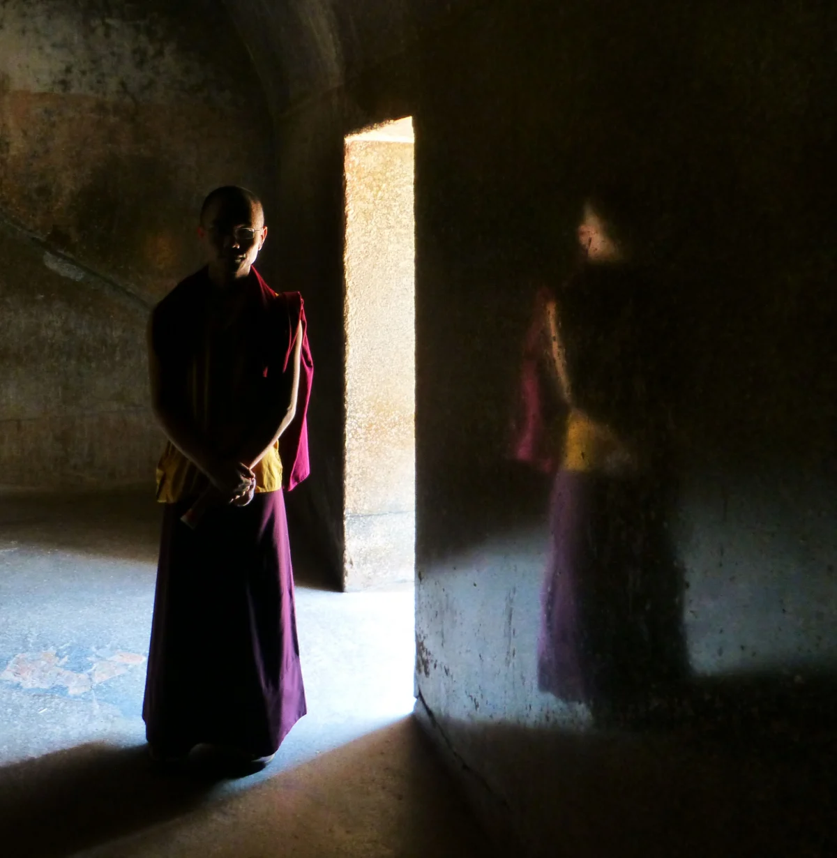 A monk in dark red robes stands in the light of a doorway inside a cave hall, and a faint image of the same figure shows in the polished wall beside him.