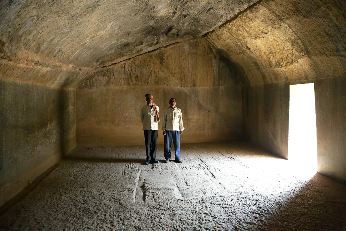 Two men stand in the Lomas Rishi cave, under a rough chiselled vault and on a rough floor, with smooth straight walls beside them.