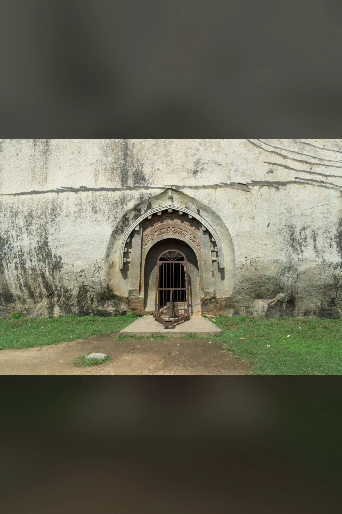 The carved arched entrance of the Lomas Rishi cave, an iron gate at its door, set in a pale wall of granite with grass in front.