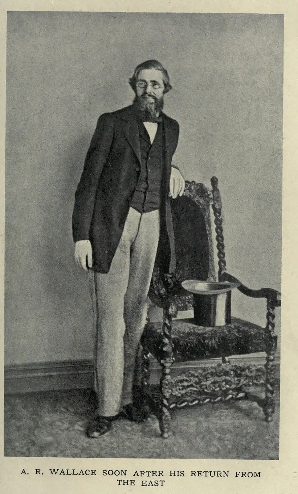 Alfred Russel Wallace in the 1860s, standing bearded beside a carved chair with a top hat on its seat.