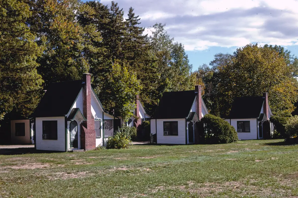 A row of small white cabins with steep dark roofs and red brick chimneys on a mown lawn, under tall trees and a partly cloudy sky.