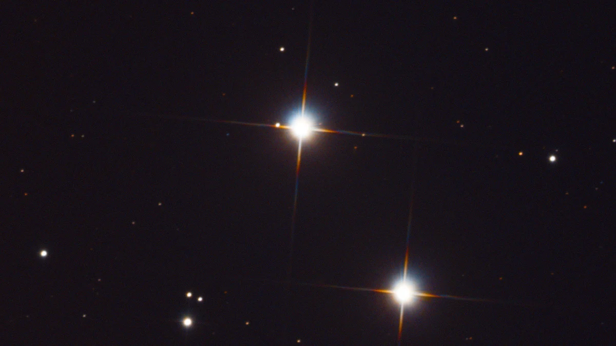 Two bright white stars with long thin diffraction spikes, one above and left of the other, on a dark field of faint orange and white points.