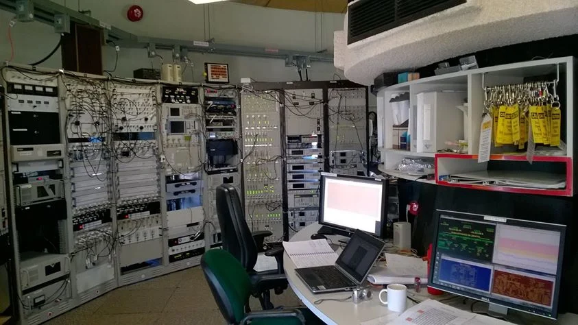 A telescope control room with tall racks of electronic equipment, a curved desk with computer monitors showing spectra, and a chair.