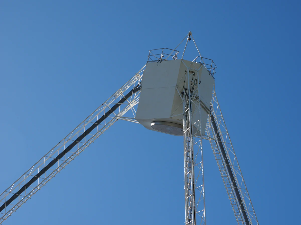 A white box-shaped cabin held high on a lattice tripod against a clear blue sky.