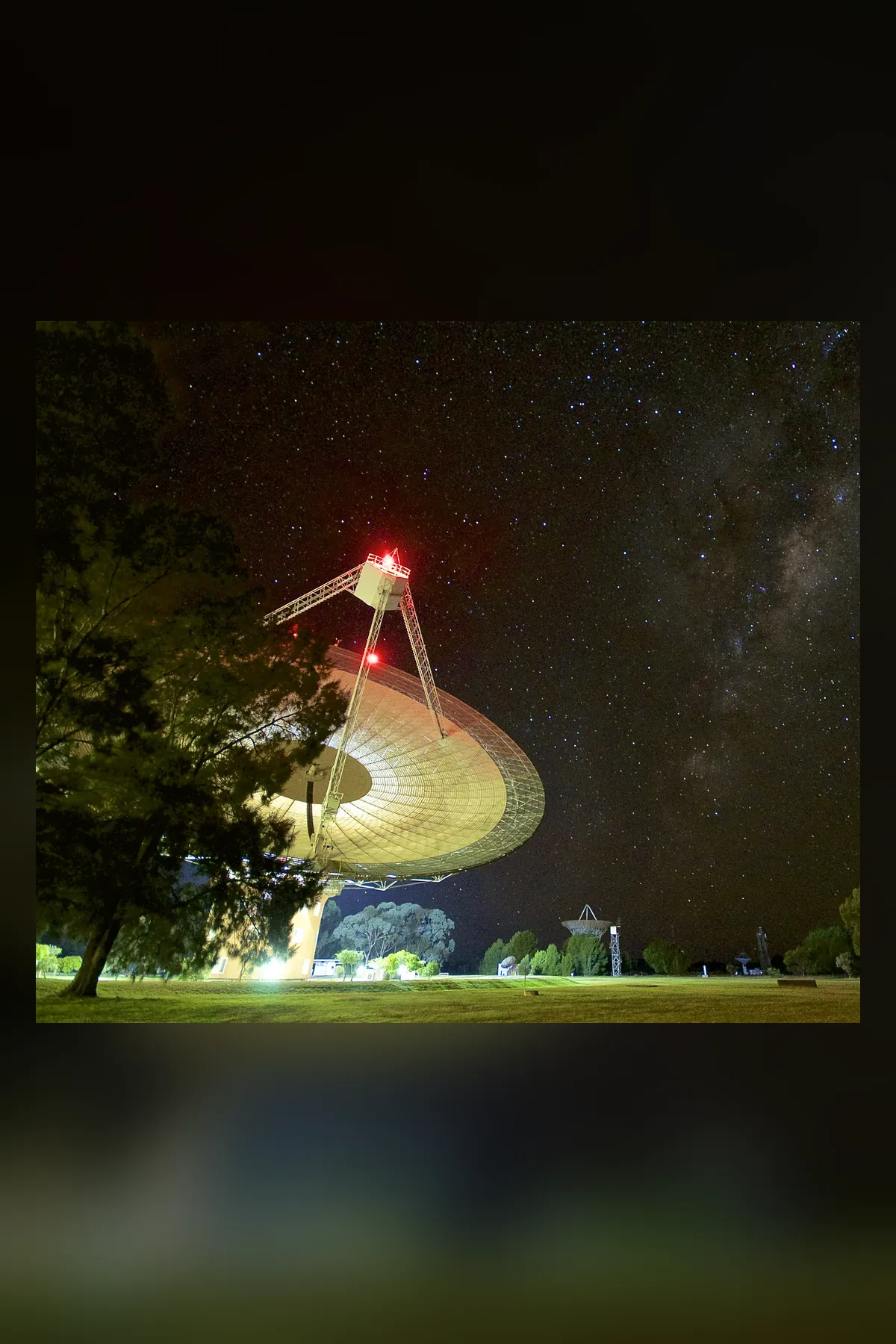 The Parkes radio telescope dish at night, tilted toward the sky, its focus cabin lit red, with the Milky Way and stars overhead and trees at the left.