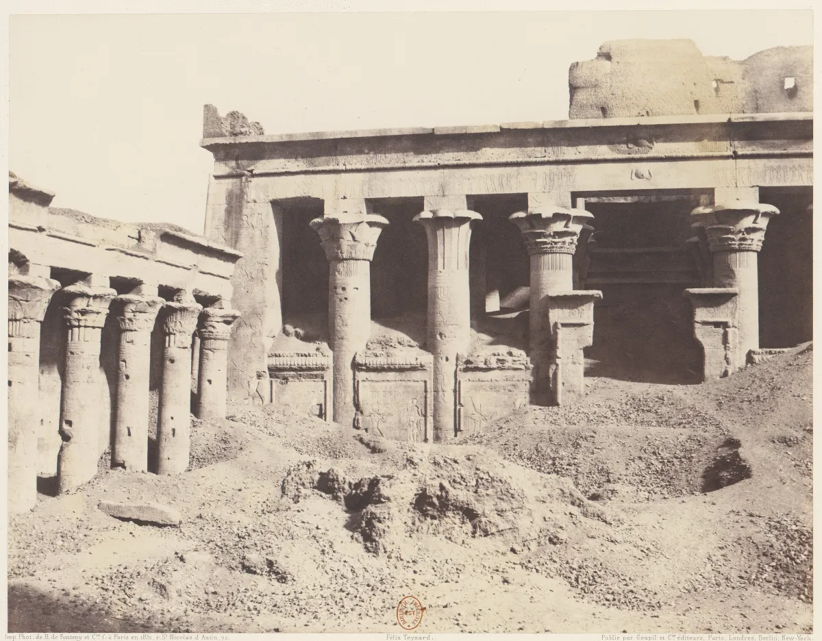 A temple court with carved columns and a wall panel half hidden behind heaps of rubble and earth.