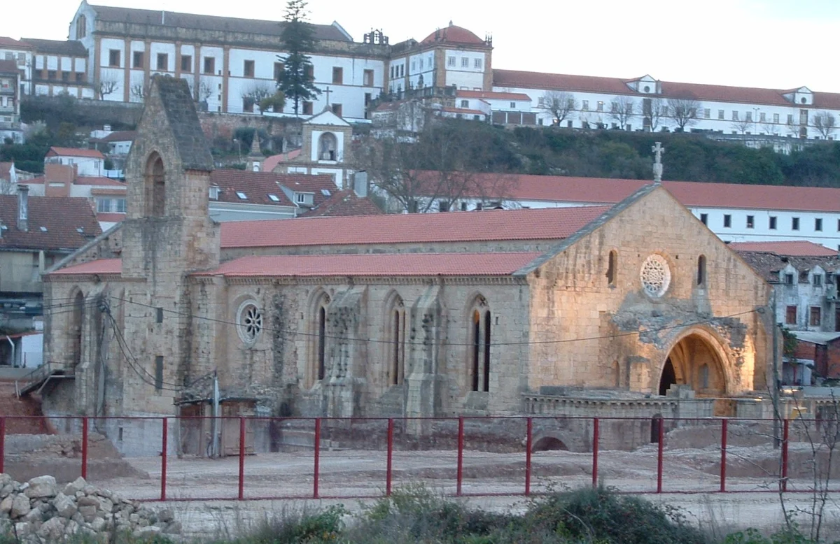 A stone Gothic church seen from the side with a red tile roof, its lower walls and a great arch exposed in a dug area behind a red fence.
