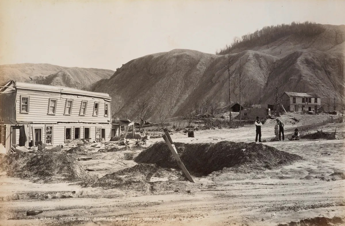 A two-storey wooden hotel with broken windows beside a heap of ash and a dug trench, three men standing on bare grey ground under bare hills.