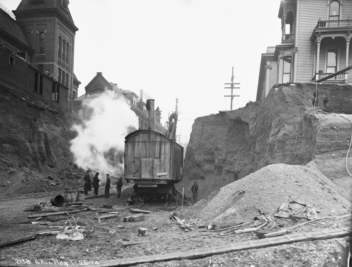 A deep street cutting between buildings with a steam excavator and workers on the exposed earth below.