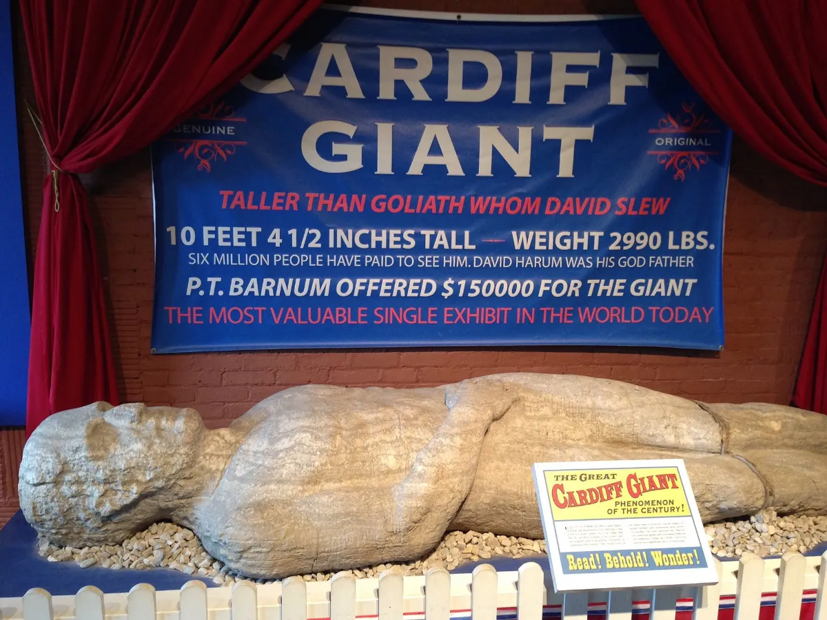 The stone figure lying on gravel in a museum barn under a large blue banner reading CARDIFF GIANT.