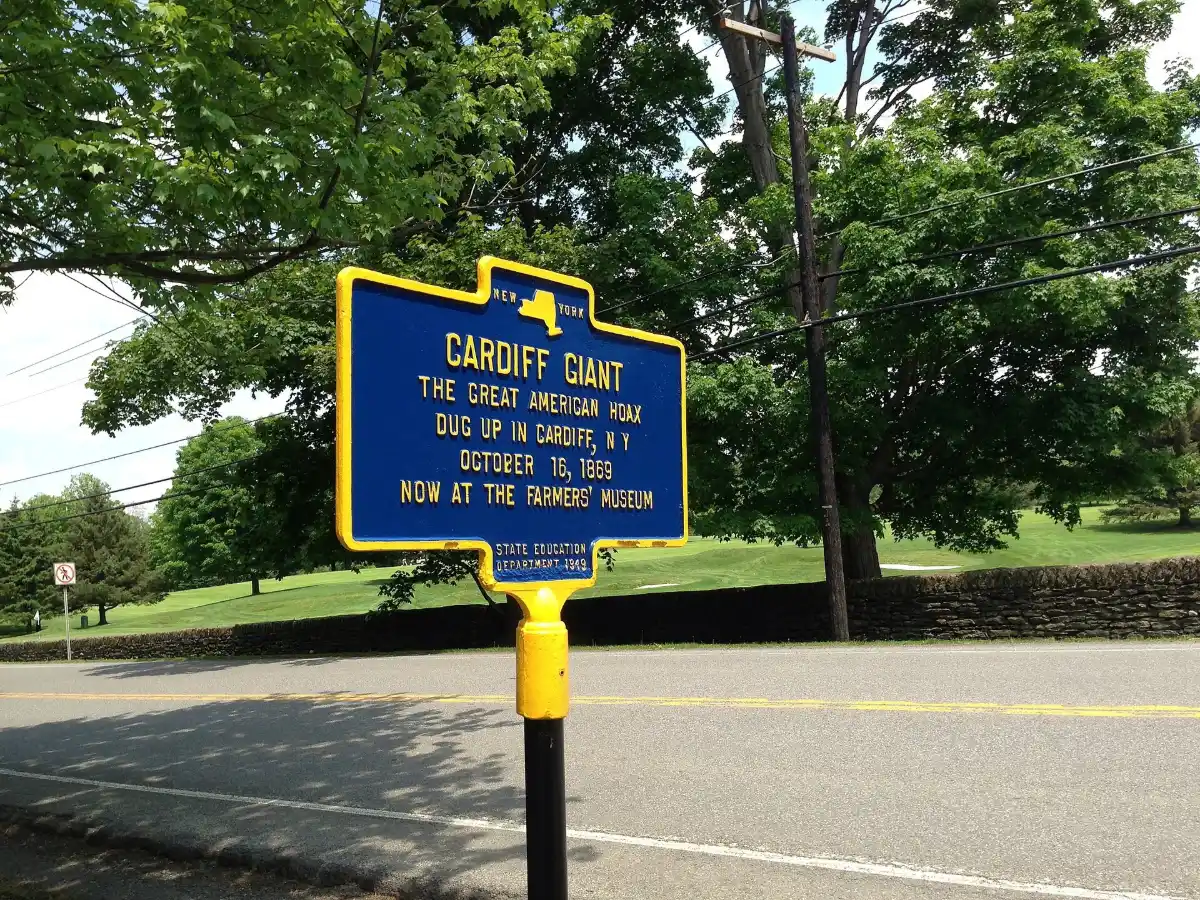 A blue and yellow roadside historic marker reading CARDIFF GIANT, beside a road under trees.