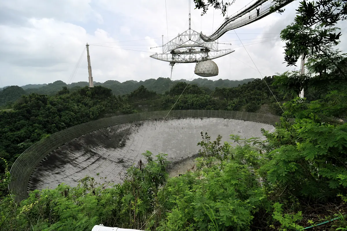 The huge Arecibo radio dish set in a green jungle valley in Puerto Rico, with its receiver platform hanging on cables above it.