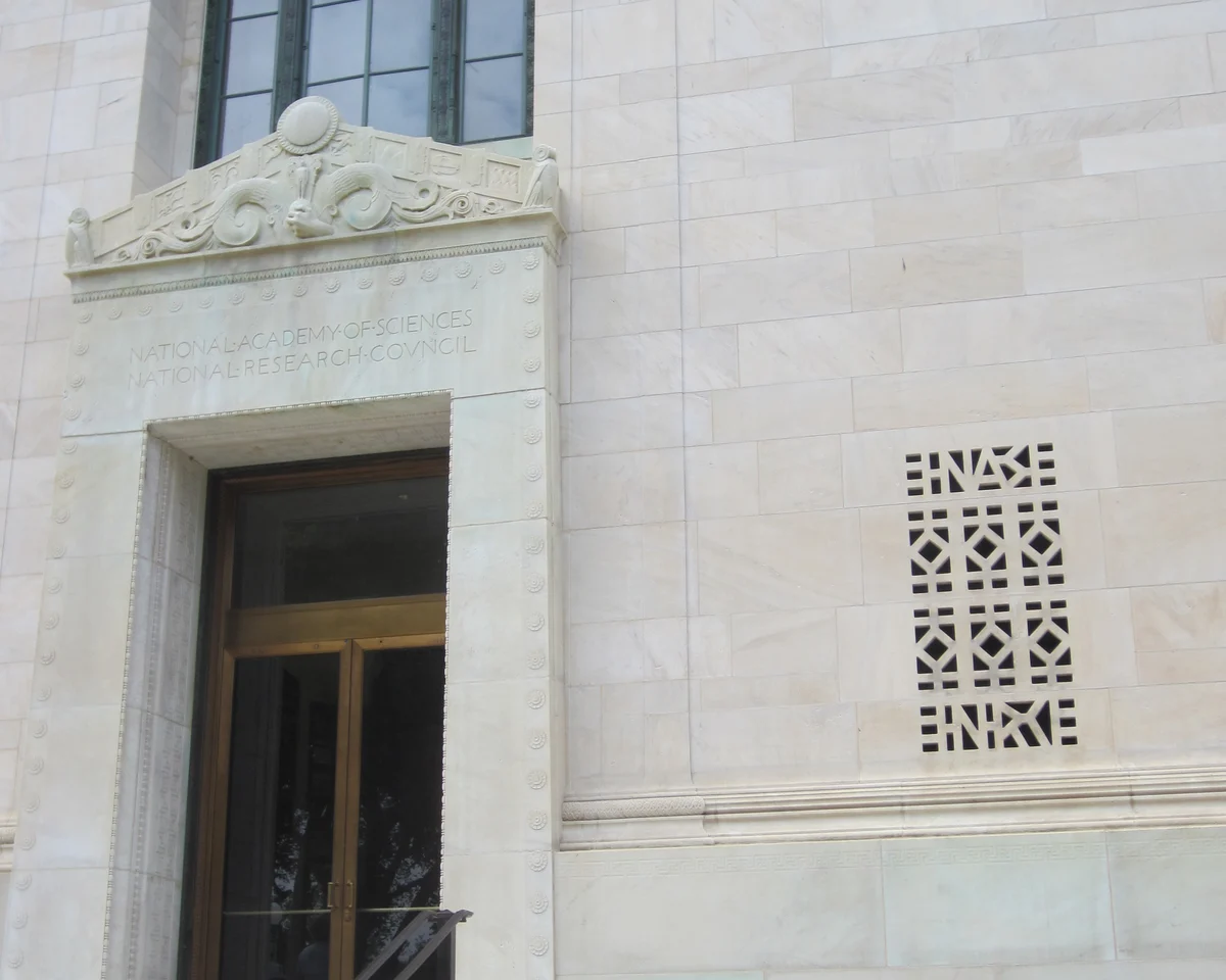 A pale marble entrance with a carved ornament above the door, an inscription reading National Academy of Sciences, a bronze double door and a pierced stone grille to the right.