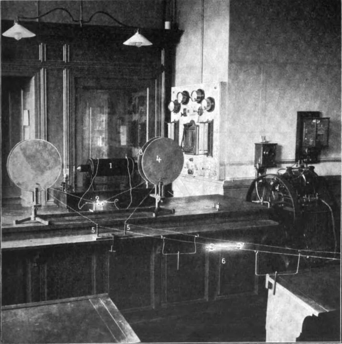 A laboratory of about 1907: two metal plates on stands, a large induction coil, a switchboard behind, and a small glowing tube bridging two long parallel wires.