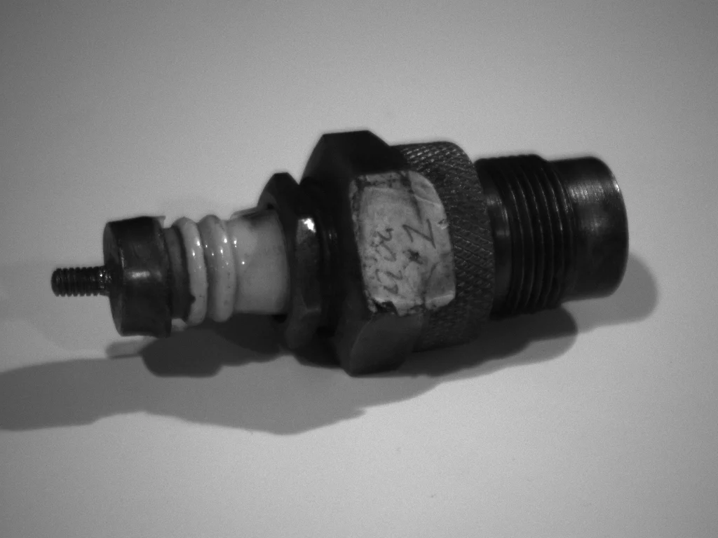 An old spark plug in black and white: a threaded metal base, a knurled body, a white porcelain insulator and a small cap at the top.