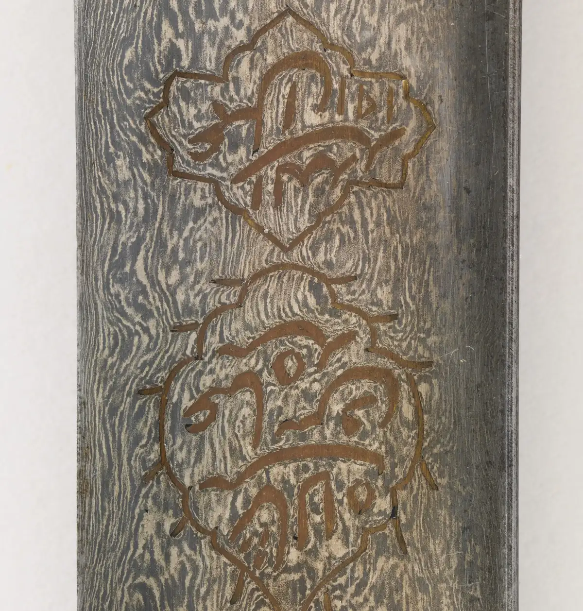 A close view of a steel blade covered in pale, wavering bands like flowing water, with two ornate cartouches of inlaid gold lettering.