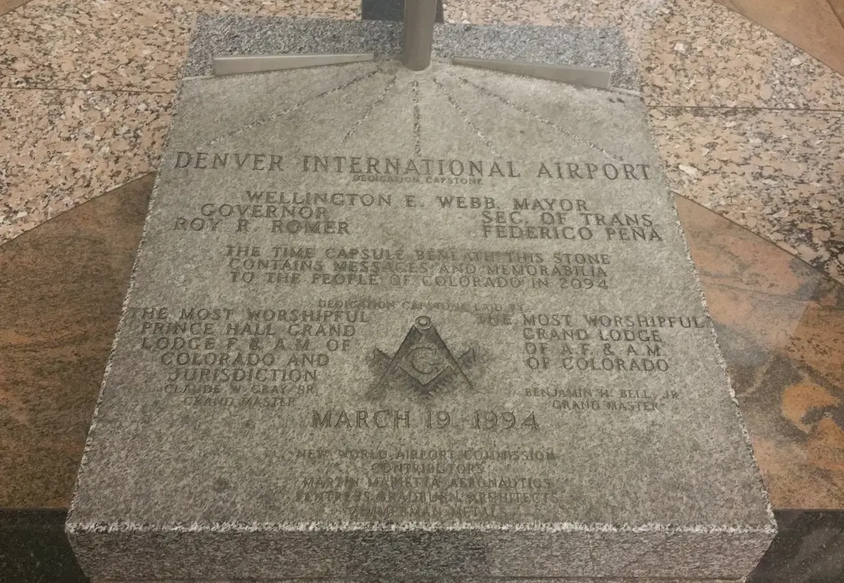 A rectangular granite slab set in a polished stone floor, cut with capital letters, a square and compasses and the date March 19 1994, with rays running to a steel post at its far edge.