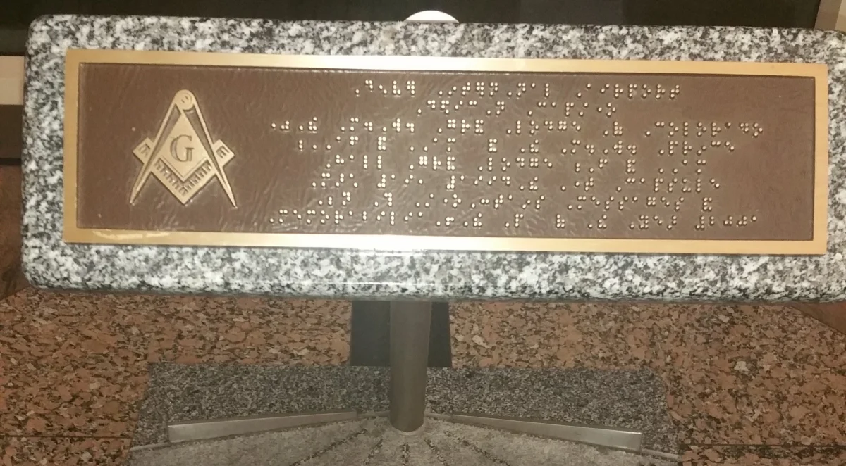 A brass plate with rows of raised dots and a square and compasses emblem, set in a speckled granite block held up on a steel post over the slab.