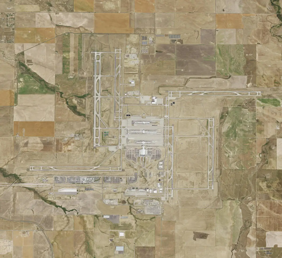 An overhead photograph of pale runways laid out in a pinwheel around three parallel concourses and a terminal, among square farm fields.