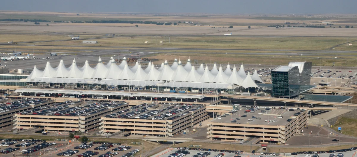 An aerial view of a long line of white peaked roofs behind wide parking garages, a dark glass hotel at right, and flat open country to the horizon.