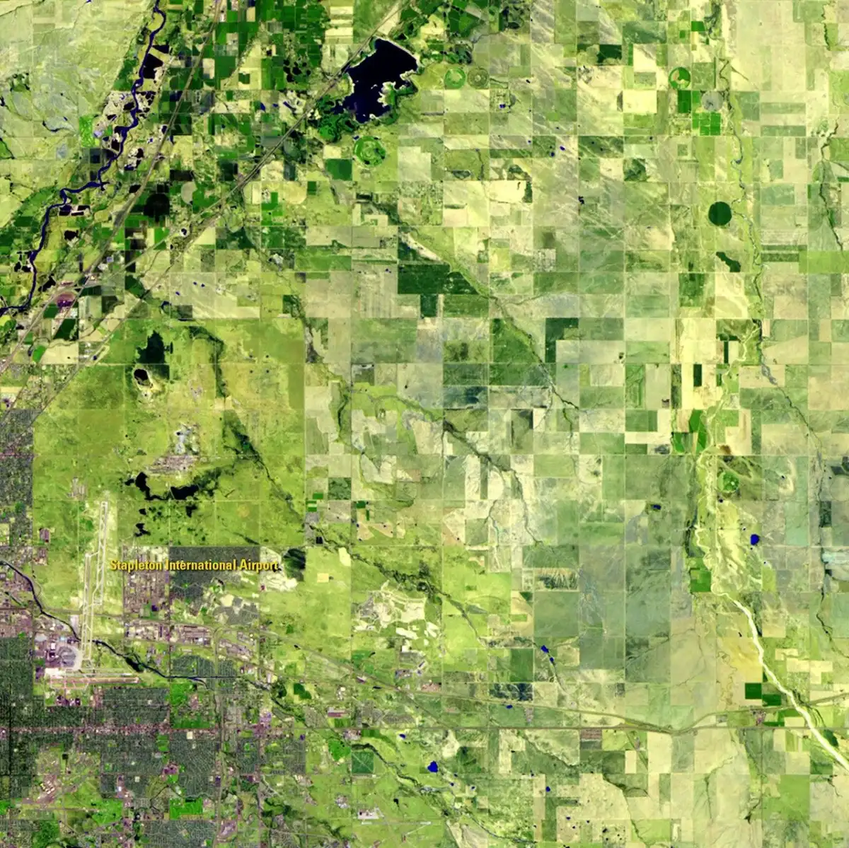 A satellite view of square green fields, with a small airport and the edge of the city at lower left.