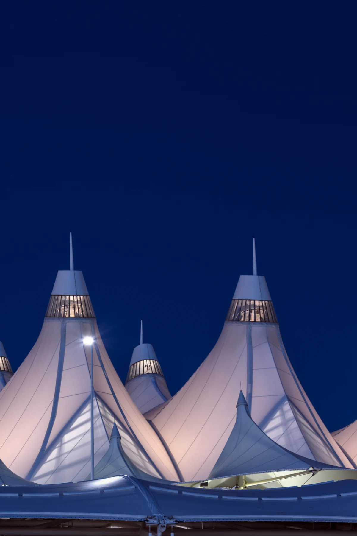 Peaked white fabric roofs of the terminal, glowing from inside, against a deep blue evening sky.