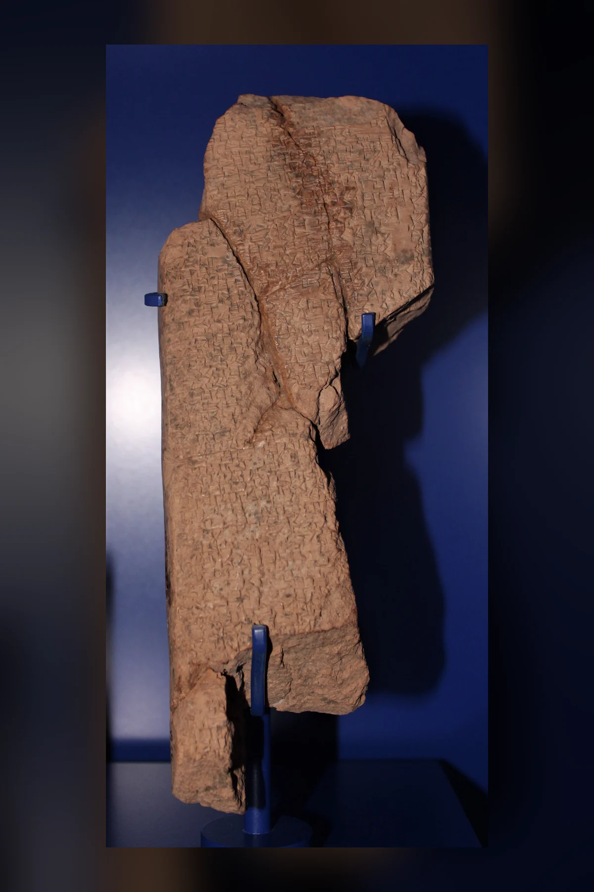 A tall, broken clay tablet fragment, light brown and covered in fine rows of cuneiform, standing on a mount against a dark blue background.