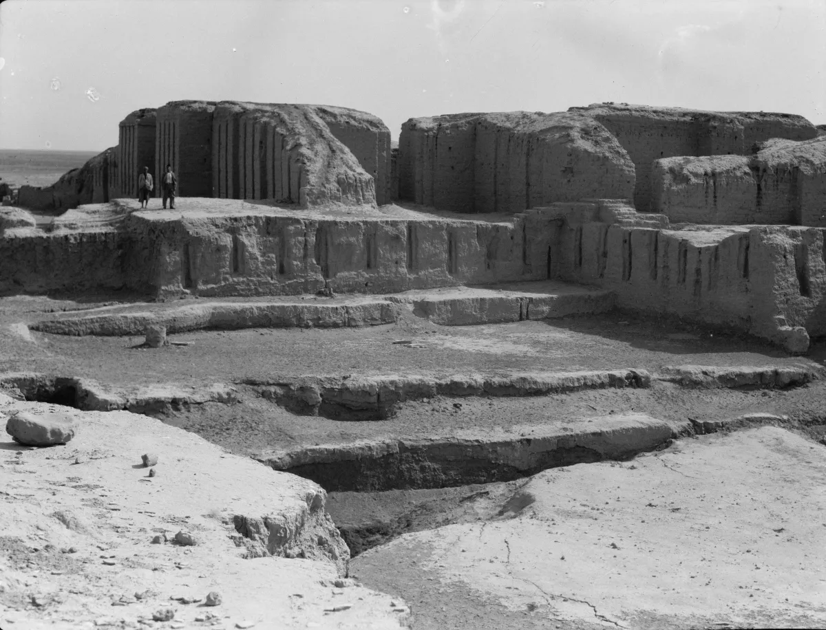 Black and white photograph of eroded mud-brick ruins on a mound in a flat desert, with two small human figures for scale.