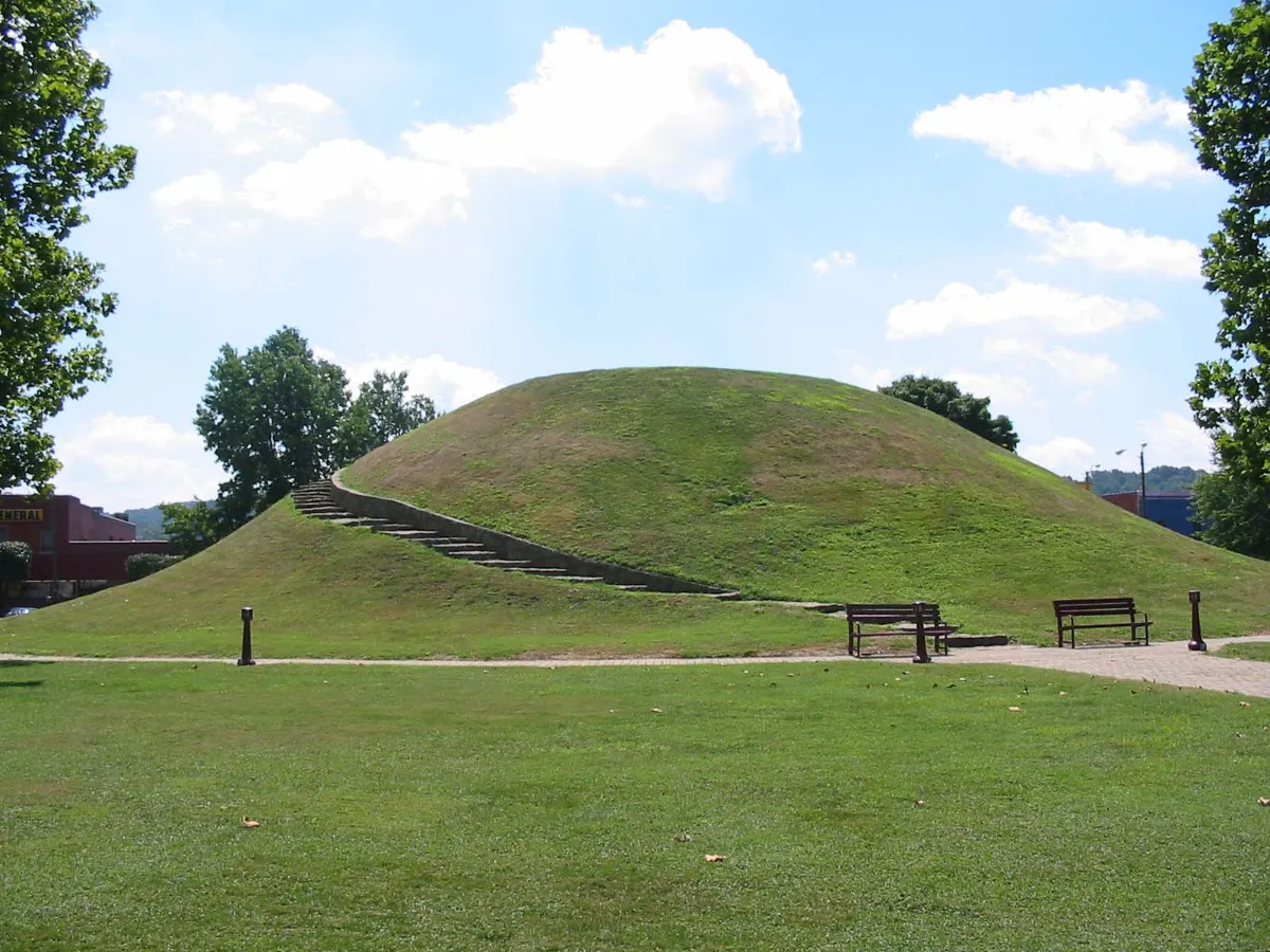 A tall grassy conical mound in a small park with trees around it.