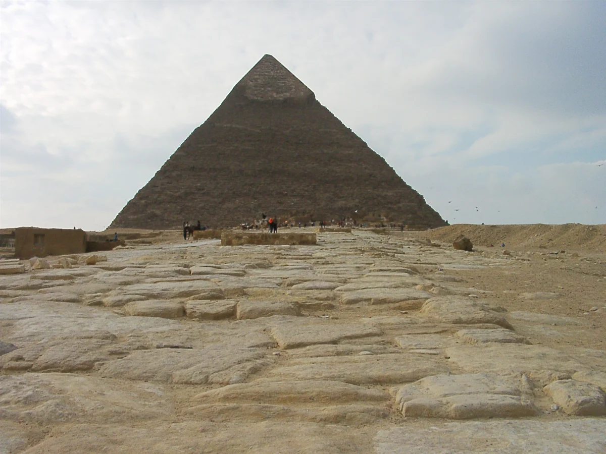 A wide stone roadway of worn blocks running straight toward the Pyramid of Khafre under a pale sky.