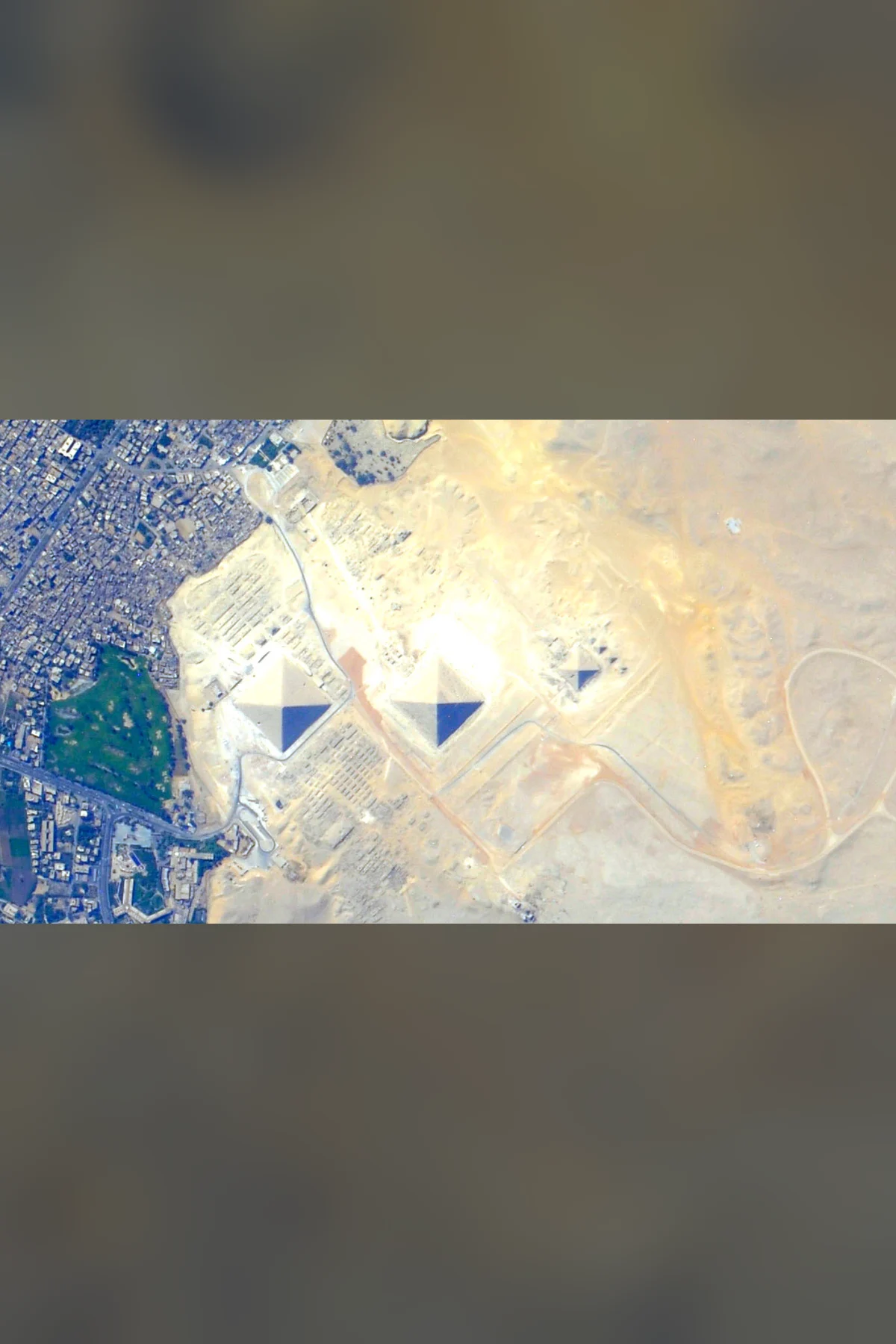 The three pyramids of Giza from straight above, in a row across a pale limestone plateau, with the city of Giza at left and the desert at right.