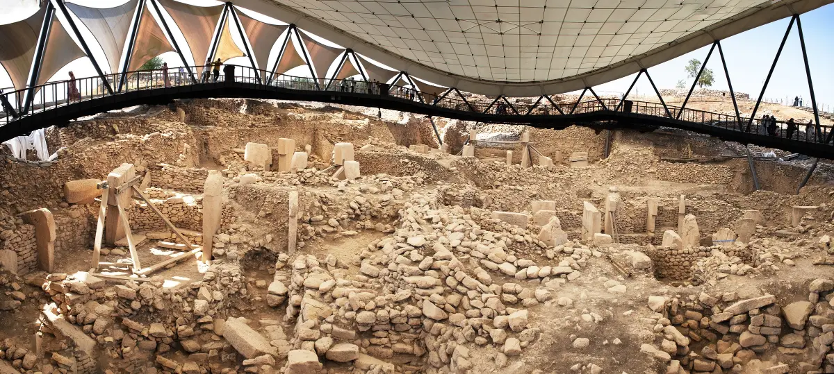 A wide view of a large excavation under a curved white roof, with rings of standing stone pillars in pits.