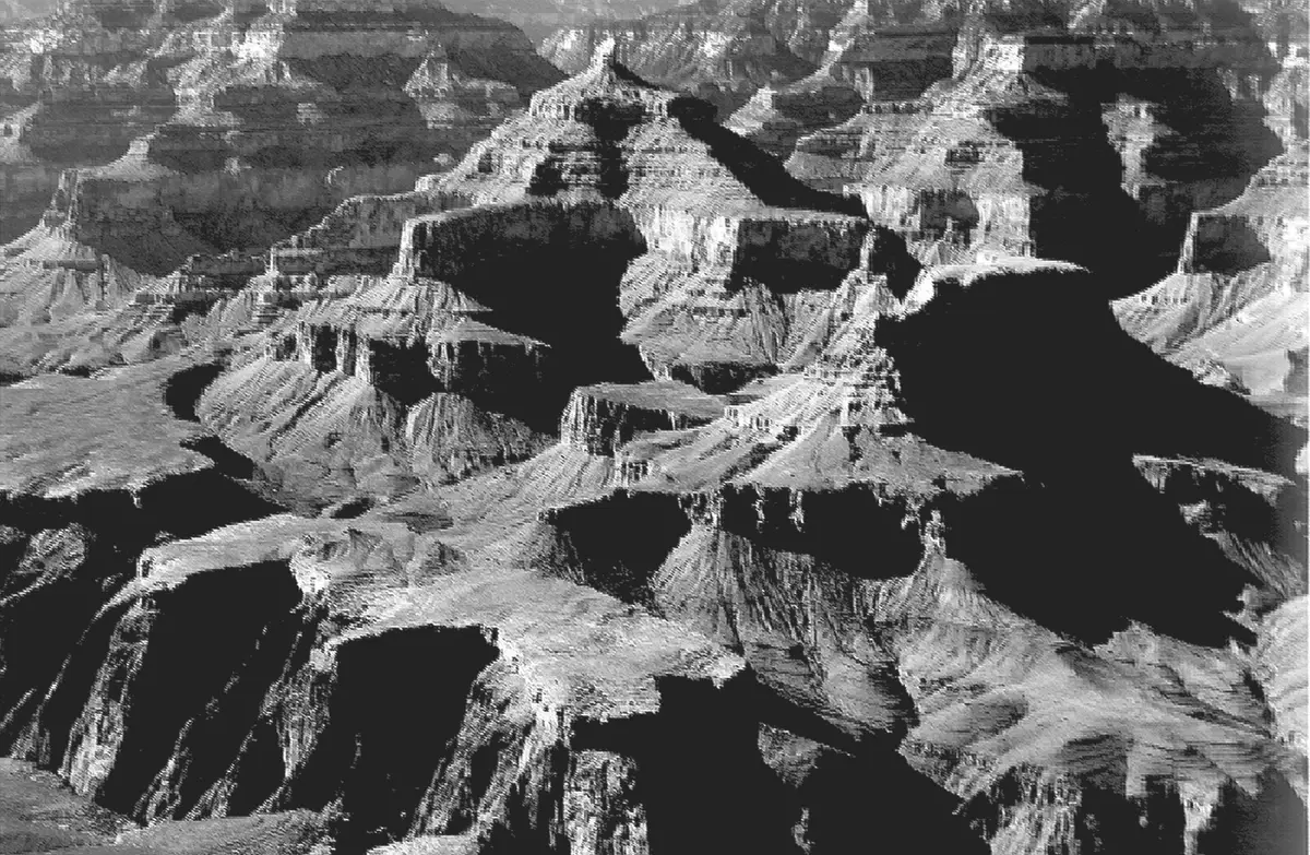A black and white photograph of stepped rock buttes in deep shadow, a flat-topped block at the centre.